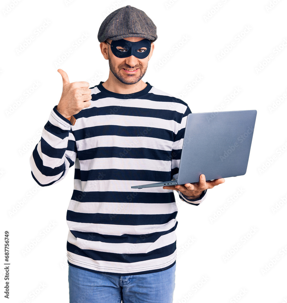 Young handsome man wearing burglar mask using laptop smiling happy and positive, thumb up doing excellent and approval sign Young handsome man wearing burglar mask using laptop smiling happy and positive, thumb up doing excellent and approval sign
