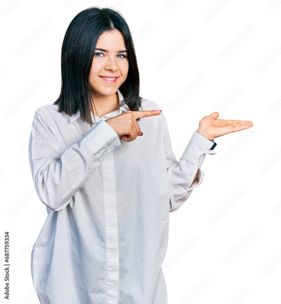 Young brunette woman with blue eyes wearing oversize white shirt amazed and smiling to the camera while presenting with hand and pointing with finger. Young brunette woman with blue eyes wearing oversize white shirt amazed and smiling to the camera while presenting with hand and pointing with finger.
