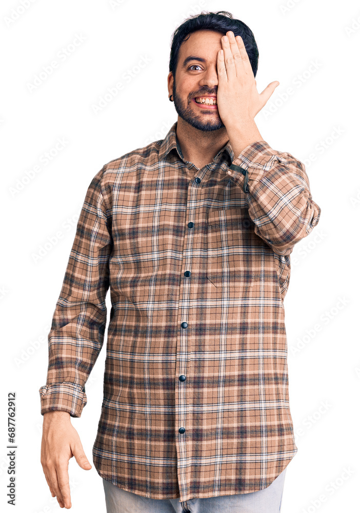 Young hispanic man wearing casual clothes covering one eye with hand, confident smile on face and surprise emotion. Young hispanic man wearing casual clothes covering one eye with hand, confident smile on face and surprise emotion.