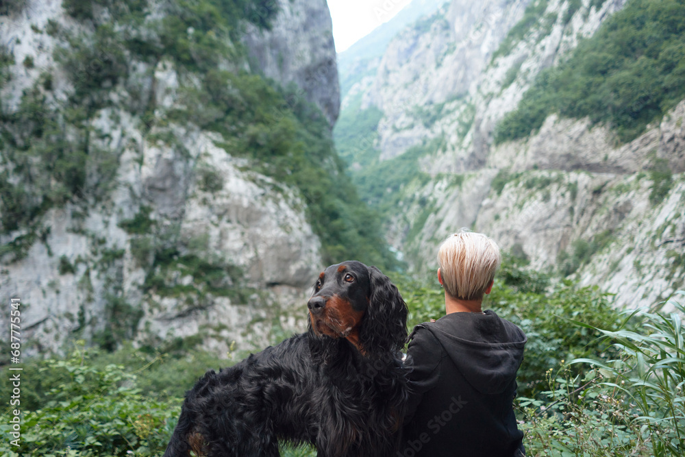 A woman and her Gordon Setter savor a tranquil moment amidst majestic cliffs. This image showcases a peaceful bond between the woman and her dog, with nature's grandeur as the backdrop