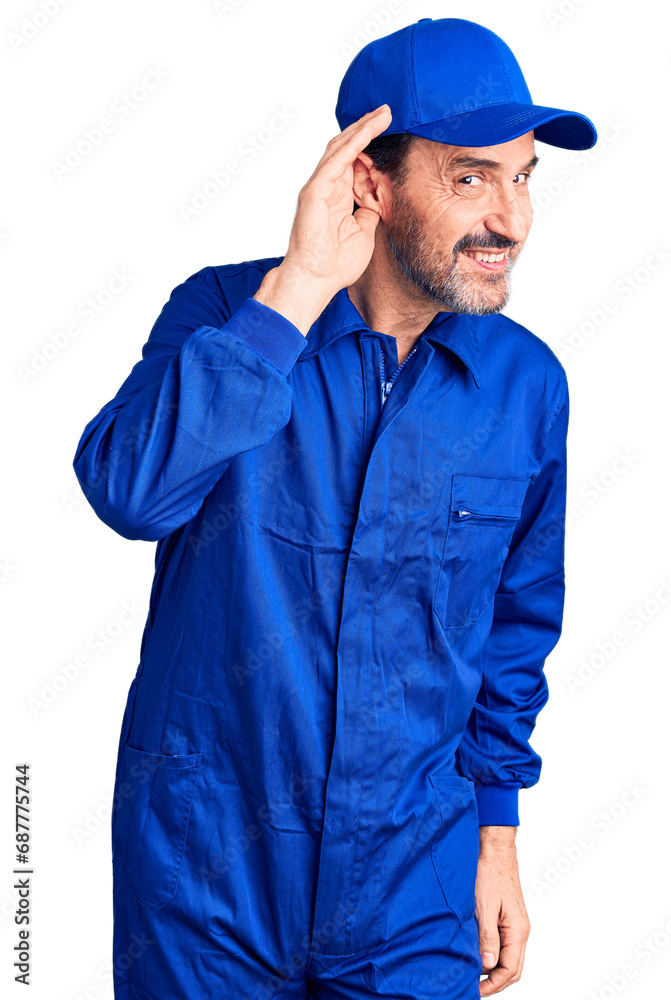 Middle age handsome man wearing mechanic uniform smiling with hand over ear listening an hearing to rumor or gossip. deafness concept. Middle age handsome man wearing mechanic uniform smiling with hand over ear listening an hearing to rumor or gossip. deafness concept.