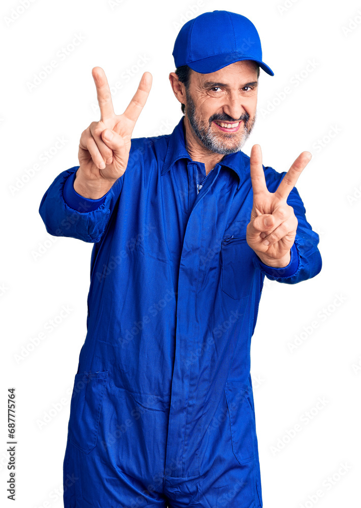 Middle age handsome man wearing mechanic uniform smiling with tongue out showing fingers of both hands doing victory sign. number two.