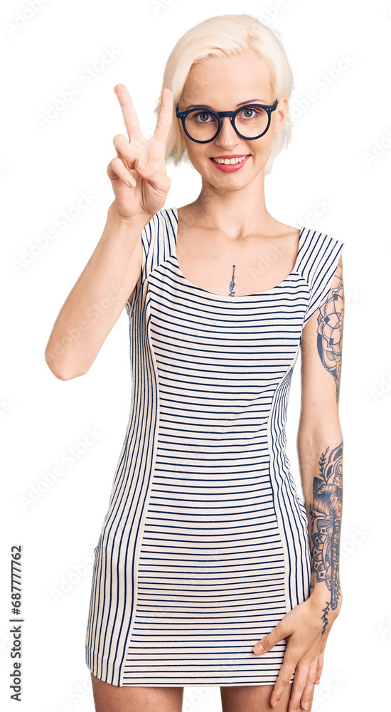 Young blonde woman with tattoo wearing casual clothes and glasses showing and pointing up with fingers number two while smiling confident and happy. Young blonde woman with tattoo wearing casual clothes and glasses showing and pointing up with fingers number two while smiling confident and happy.