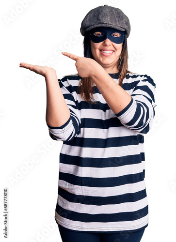 Young beautiful brunette woman wearing burglar mask amazed and smiling to the camera while presenting with hand and pointing with finger. © Krakenimages.com