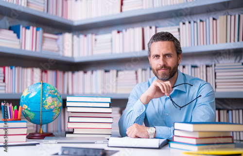 Portrait of teacher with book in library classroom. Handsome teacher in university library. Teachers Day. Teacher giving classes. School teacher in library. Tutor at college library on bookshelf.