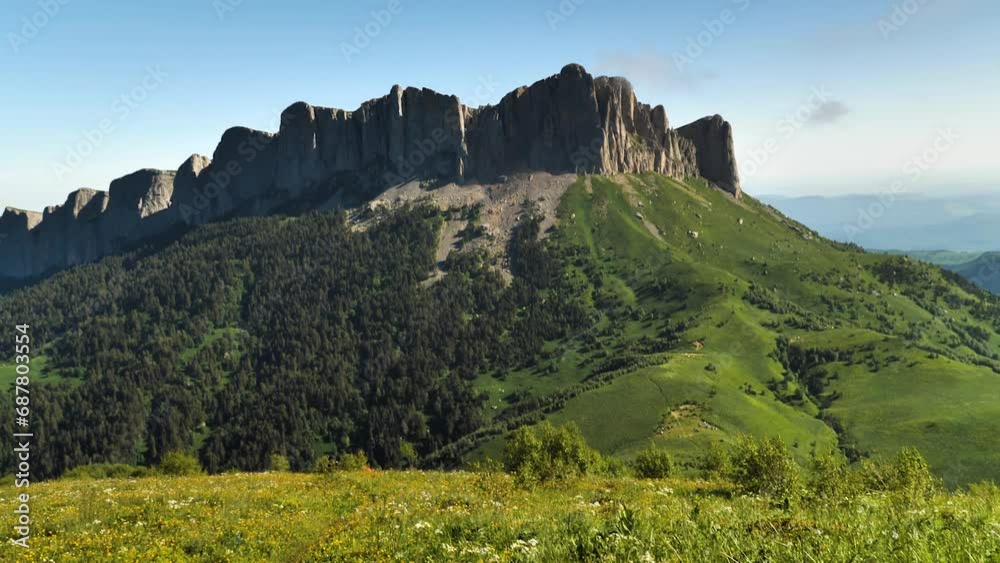 A high mountain with a long sheer rock wall. Blue sky in the background. Trees and grass grow on the mountainside. Panorama from bottom to top. The beauty of nature of the Caucasus Mountains A high mountain with a long sheer rock wall. Blue sky in the background. Trees and grass grow on the mountainside. Panorama from bottom to top. The beauty of nature of the Caucasus Mountains