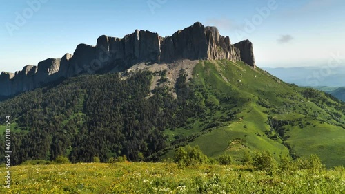 A high mountain with a long sheer rock wall. Blue sky in the background. Trees and grass grow on the mountainside. Panorama from bottom to top. The beauty of nature of the Caucasus Mountains photo