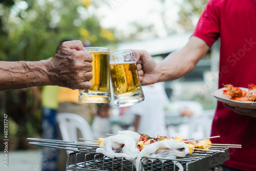 Outdoor barbecue with various meats on grill, two people holding plates and drinks, smoke rising, festive and casual atmosphere. holding glass of beer. family party. photo