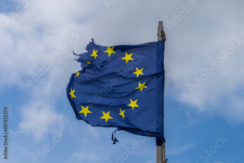A torn EU flag blows in the wind with a few clouds in the background
