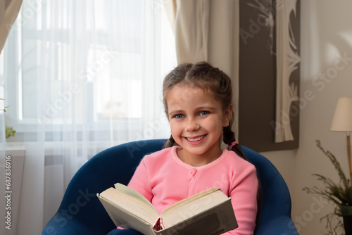girl reading a book. a little girl with pigtails in a pink jacket sits on a blue chair and reads a thick book in a white cover