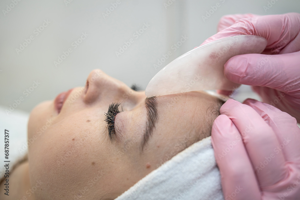Naklejka premium cosmetologist masseur doing faacial anti-agging procedure with gouache scraper for her client