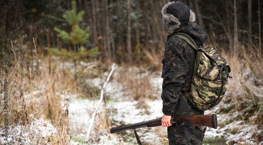 Male hunter in camouflage and with backpack, armed with a rifle, walks through the snowy winter forest