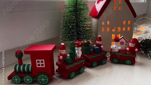 Christmas decorations are on the windowsill in winter. A candlestick house, an artificial small Christmas tree, a red toy train, a fir branch and a garland lie on a white windowsill. A Christmas card