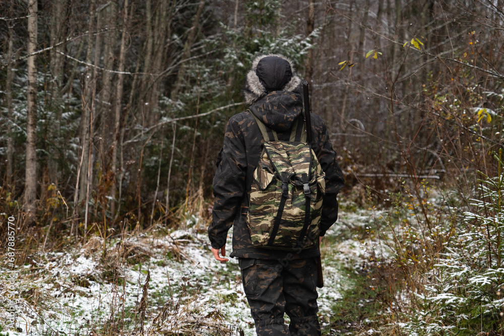 Male hunter in camouflage and with backpack, armed with a rifle, walks through the snowy winter forest Male hunter in camouflage and with backpack, armed with a rifle, walks through the snowy winter forest