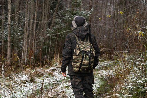Male hunter in camouflage and with backpack  armed with a rifle  walks through the snowy winter forest
