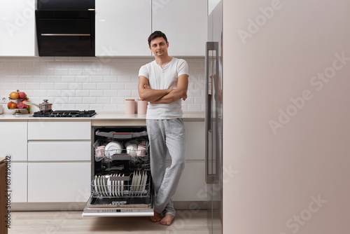 handsome positive man using dishwasher and washing dishes in white modern kitchen. copy space