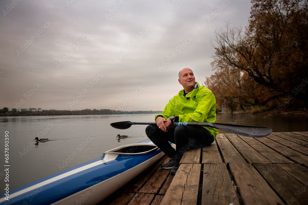 A middle-aged man sitting on the jetty and ready to go kayaking on the river