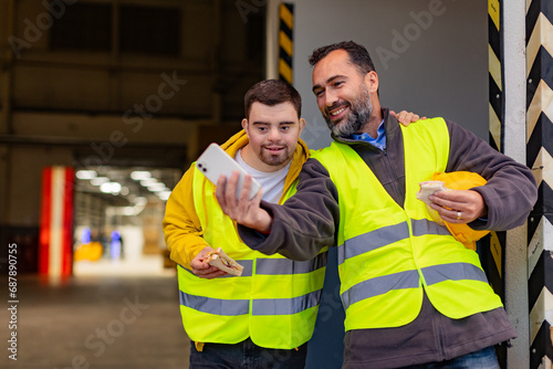 Man with Down syndrome and his colleague taking break from work, taking selfie on smartphone, having fun. Concept of workers with disabilities, support in workplace.