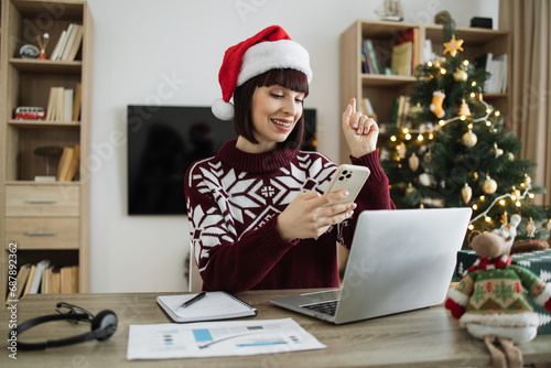 Positive female in Santa hat holding smartphone raise finger up, sign of good idea while staying in workplace at background of Christmas tree at home.