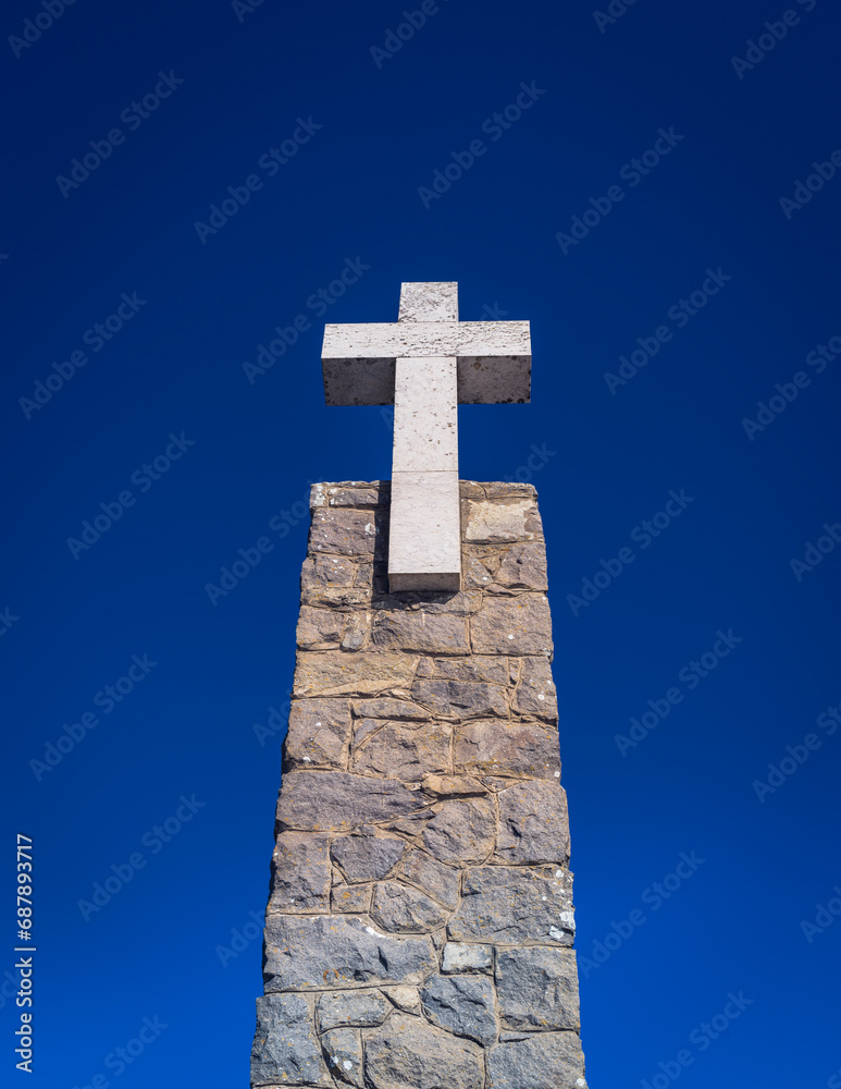 A column made of stones with a white cross at the end; bottom view on blue sky background.