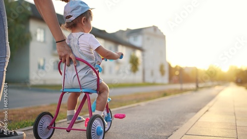 mother teaches daughter to ride. happy family kids dream concept. young mother teaches her daughter a small tricycle along city center road. young mother teaches her little daughter ride bicycle