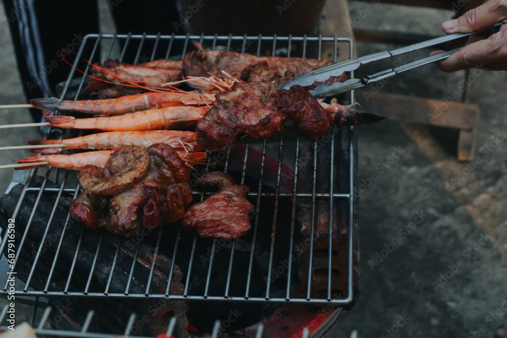 People are cooking seafood and vegetable skewers on a charcoal grill. with a plate of shrimp placed next to it A warm family holiday celebration. BBQ party People are cooking seafood and vegetable skewers on a charcoal grill. with a plate of shrimp placed next to it A warm family holiday celebration. BBQ party
