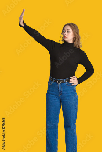 Cute young Caucasian woman with a serious expression on her face and a hand gesture indicating refusal. Full height. Yellow background.