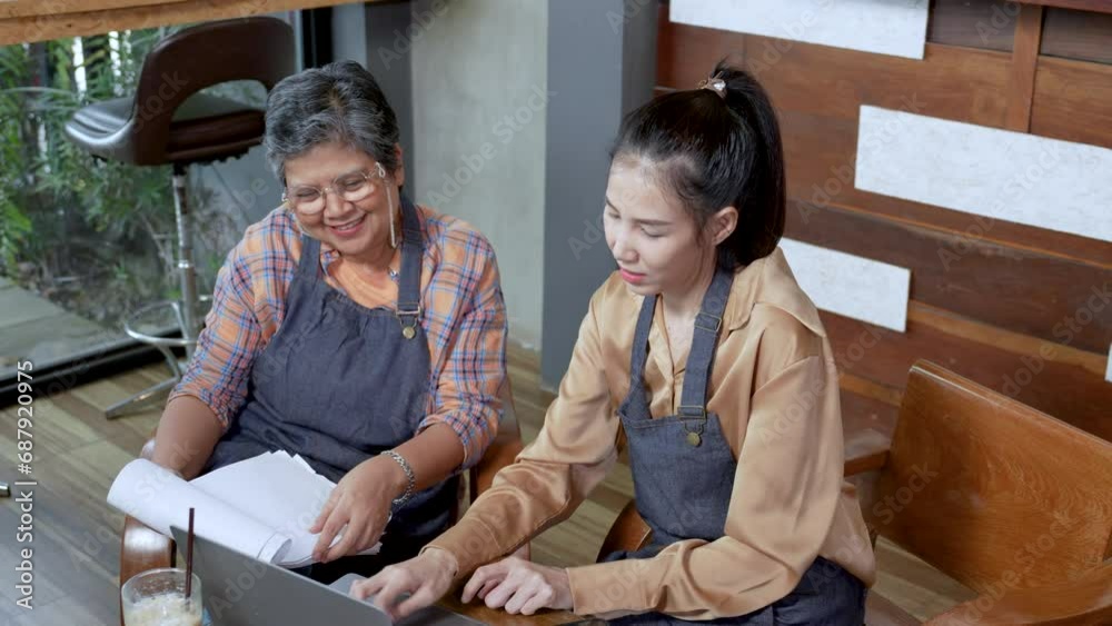 Elderly coffee shop owner in apron smiling working on laptop on table with daughter. Senior shop owner drinking iced coffee and do account and online stuff on computer. Staff working on accounting. Elderly coffee shop owner in apron smiling working on laptop on table with daughter. Senior shop owner drinking iced coffee and do account and online stuff on computer. Staff working on accounting.
