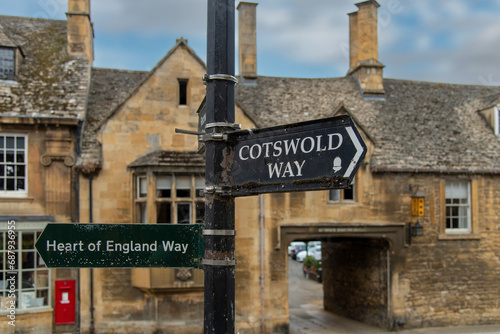 Close up of the signage in the middle of Chipping Campden, UK indicating the Heart of England Way and Cotswold Way for hiking with out of focus typical Cotswolds limestone cottage in background photo