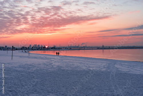 Fototapeta Naklejka Na Ścianę i Meble -  Beautiful sunset on the coast in winter. White snow lies on the embankment. Winter city beach. Amazing clouds sky horizon. Baltic Sea. Tallinn silhouette. Bright golden orange pink purple tones.