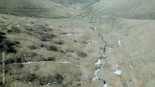 Aerial view of the valley of a winding small mountain river. Snow thawed along the banks, trees without leaves. Deserted gray mountains in winter without grass. Drone footage. Direct flight. Armenia