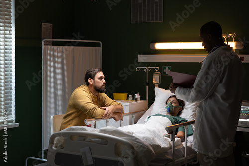 Man stays by the unconscious girl's side in the hospital and African doctor stands near woman and writes down notes.