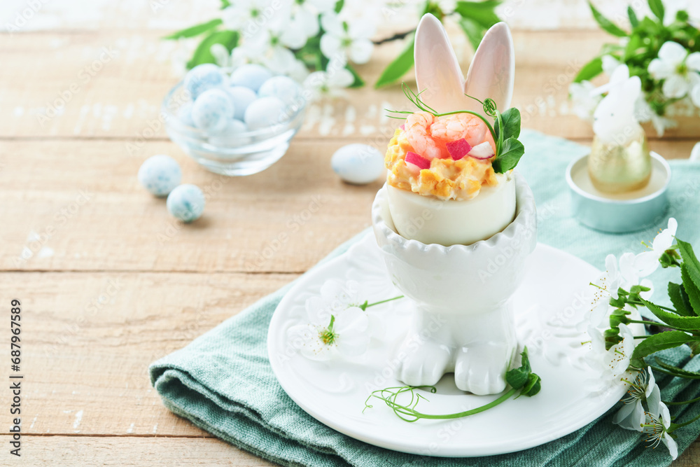 Stuffed or deviled eggs with yolk, shrimp, pea microgreens with paprika in rabbit-shaped stand for easter table decorate fresh cherry or apple blossoms on light background. Traditional dish for Easter Stuffed or deviled eggs with yolk, shrimp, pea microgreens with paprika in rabbit-shaped stand for easter table decorate fresh cherry or apple blossoms on light background. Traditional dish for Easter