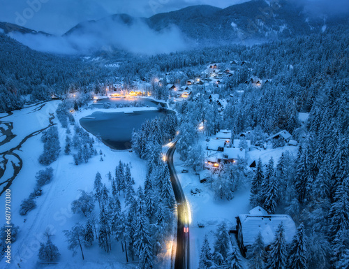 Aerial view of fairy alpine village in snow, road, forest, Jasna lake, houses, street lights at winter night. Top view of mountains, illumination, snowy pine trees at twilight. Kranjska Gora, Slovenia