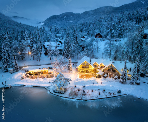 Aerial view of illuminated houses in fairy village in snow, forest, Jasna lake, street lights at winter night. Top view of alpine mountains in fog, snowy pine trees at dusk. Kranjska Gora, Slovenia photo