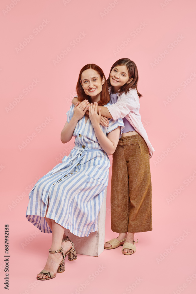 happy teenage girl embracing stylish mother sitting on stone block on pink backdrop, love and unity