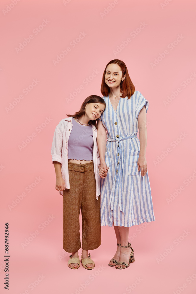 full length of cheerful woman with teen daughter holding hands and smiling at camera on pink full length of cheerful woman with teen daughter holding hands and smiling at camera on pink