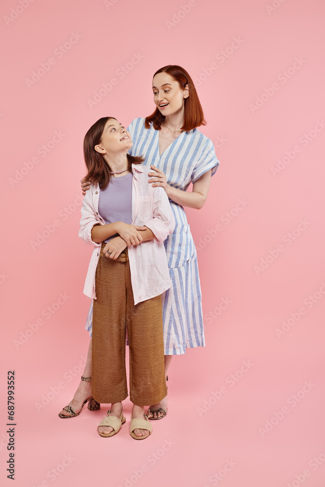 full length of happy and stylish mother with teenage daughter looking at each other on pink