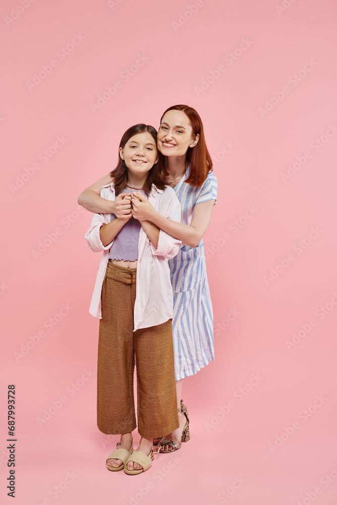 joyful woman embracing teenage daughter and holding her hands on pink backdrop, full length joyful woman embracing teenage daughter and holding her hands on pink backdrop, full length