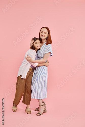 full length of stylish and joyful woman with teen daughter embracing on pink backdrop, happy family