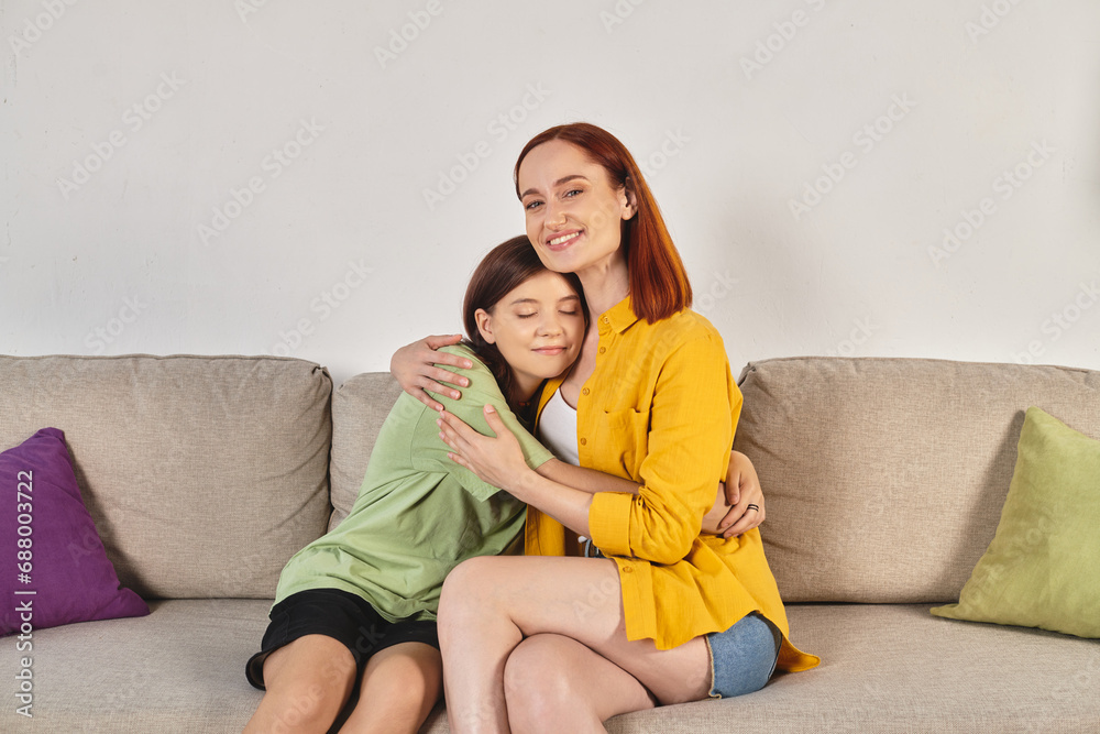 cheerful woman with teenage daughter sitting and embracing on cozy couch in modern living room cheerful woman with teenage daughter sitting and embracing on cozy couch in modern living room