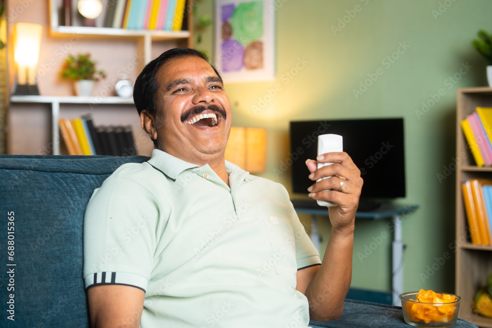 middle aged man laughing while watching comedy show on tv or television by holding remote at home - concept of entertainment, retirement lifestyle and relaxation or refreshment.