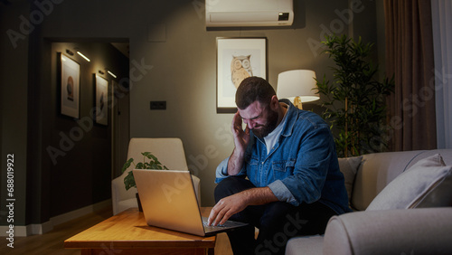 Bearded young man sitting on sofa at home while using laptop having headache. Tired young man get having pain from a headache or migraine © mahirkart