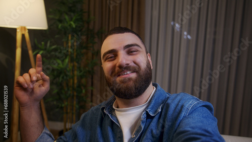 Close up young adult man holding a smart phone in hand making video call while sitting on a sofa in living room at home at night, smiling at camera, having fun, leisure