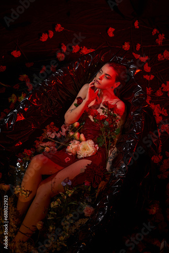 dreamy young woman in wet slip dress sitting in black bathtub with blooming flowers, red light