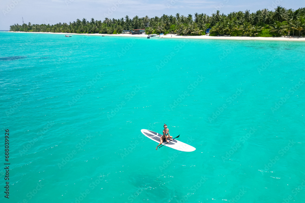 Aerial view of a woman on a white supboard in the turquoise waters of the Maldives - obrazy, fototapety, plakaty 