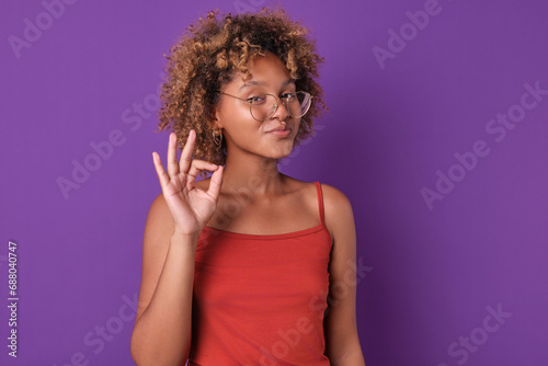 Young beautiful confident African American woman student with glasses shows confirming gesture saying that everything is ok or agreeing to your commercial proposal stands posing on purple background. photo