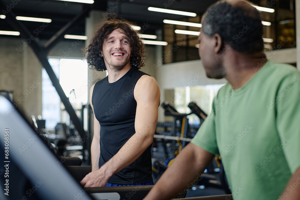 Caucasian guy and african american man looking at each other walking on treadmill at gym Caucasian guy and african american man looking at each other walking on treadmill at gym