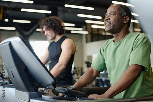 African american man walking on treadmill at gym with caucasian sportsman training on background