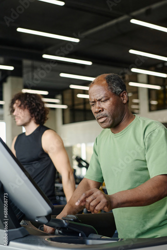 Black man about to press key on treadmill at gym with caucasian man training on background